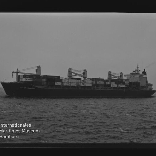Schwarzweißaufnahme eines Containerschiffs mit Ladekränen und gestapelten Containern auf See, Museumsstempel links unten.