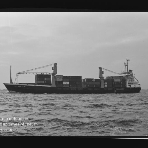 Containerfrachter mit aufgestapelten Ladecontainern und zwei an Deck montierten Kränen, schwarzweiße Aufnahme mit Museumslogo