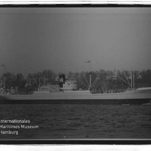 Frachtschiff in Seitenansicht auf See, mit Ladekränen und markantem Schornstein mit den Buchstaben KG