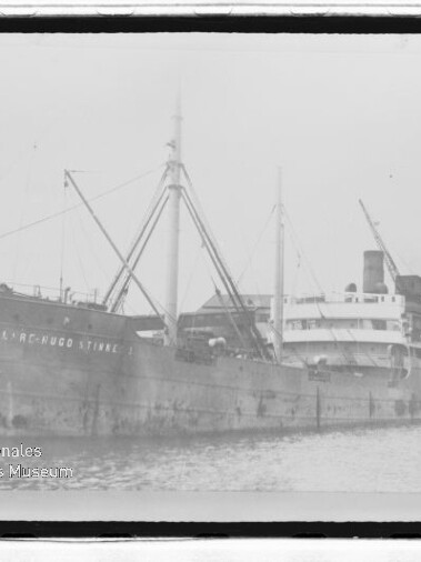 Altes Frachtschiff mit Ladebäumen und Schornstein am Kai; Name am Rumpf teilweise lesbar, Wasserzeichen des Internationalen Maritimen Museums Hamburg