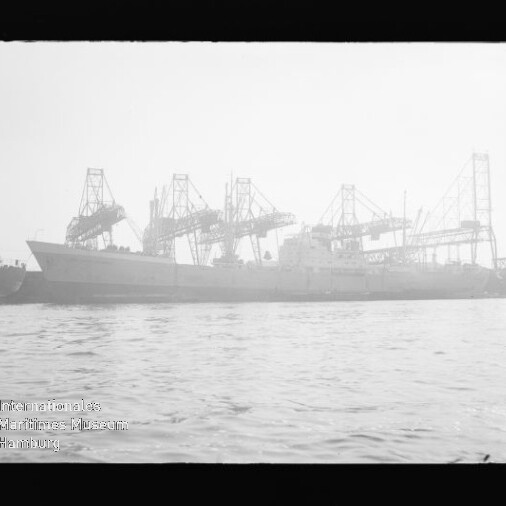 Großes Frachtschiff an Kai vor mehreren Hafenkränen bei diesigem Licht, Wasser im Vordergrund und Museumsstempel links unten.