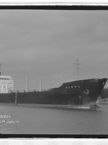 Frachtschiff Algol mit dunklem Rumpf und hellem Aufbau fährt auf ruhigem Kanal, Baumreihe am Ufer; historische Schwarzweißaufnahme.