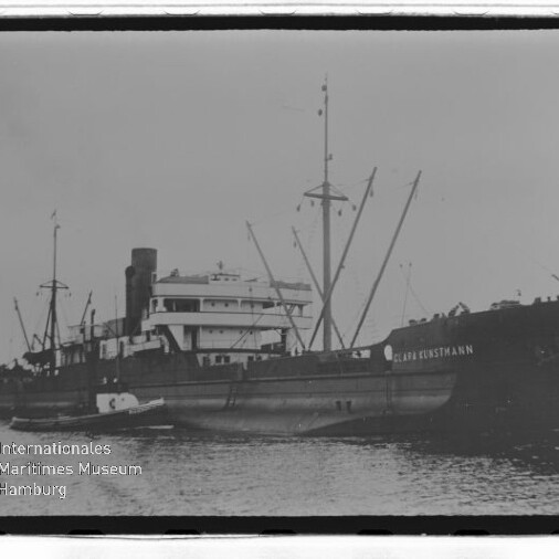 Schwarzweißfoto eines Frachtschiffs mit Schornstein und mehreren Masten, einem Beiboot am Rumpf und Museumslogo links unten.