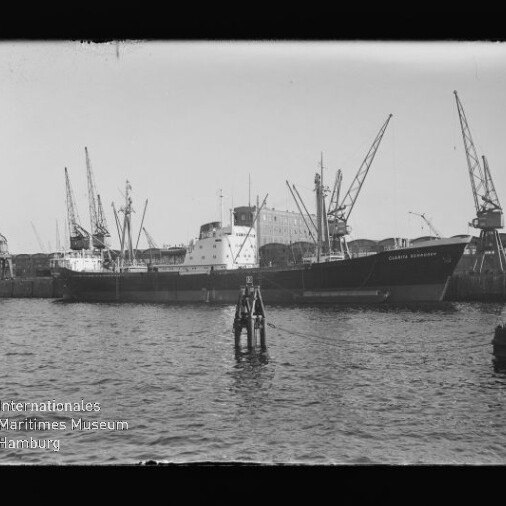 Frachtschiff am Kai mit mehreren Hafenkränen im Hintergrund und einer Boje im Wasser, Wasserzeichen des Internationalen Maritimen Museums Hamburg.