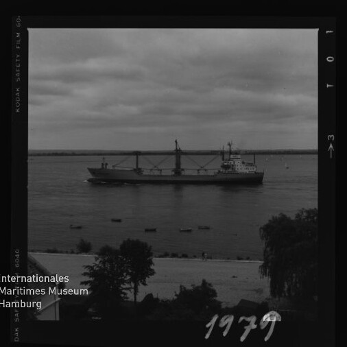 Schwarzweiß-Archivfoto (1979) eines Frachtschiffs mit Ladekränen auf ruhigem Wasser, im Vordergrund Uferbäume.