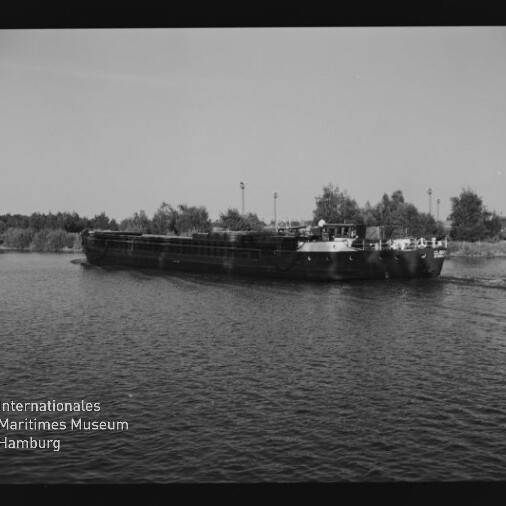 Schwarzweißfoto eines langen Binnenschiffs, das auf einem Fluss vor einem baumbestandenen Ufer vorbeifährt.