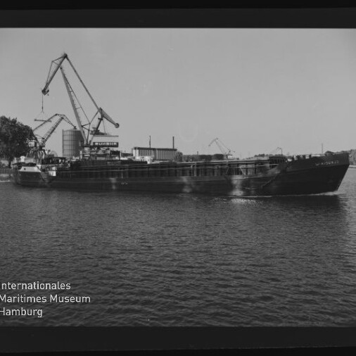Frachtschiff auf ruhigem Wasser vor einem Industriehafen mit mehreren Hafenkränen; schwarz-weißes Bild mit Museumswasserzeichen