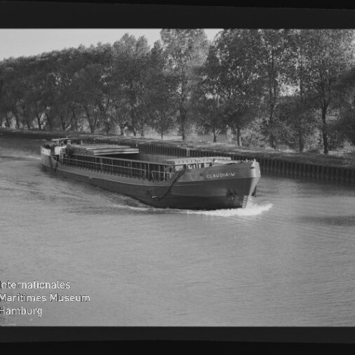 Binnenschiff Claudia fährt auf einem Kanal, Ufer mit einer Baumreihe im Hintergrund.
