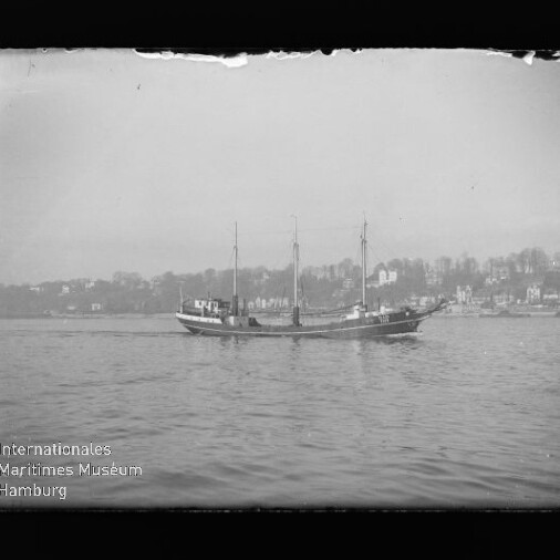 Dreimastiges Segelschiff ohne Segel fährt vor ruhiger Küste mit Häusern; Schriftzug Internationales Maritimes Museum Hamburg sichtbar.