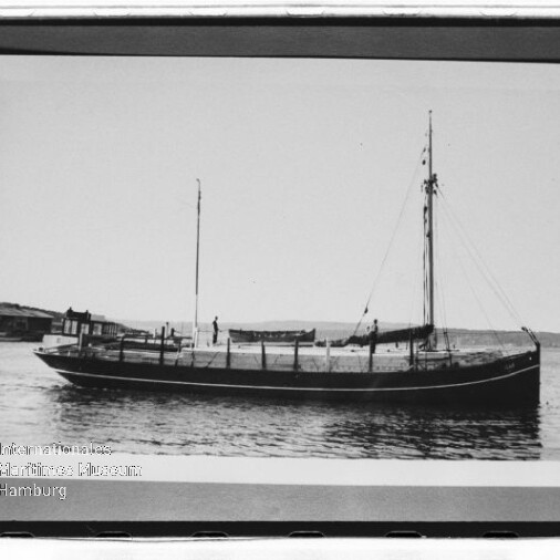 Schwarzweißfoto eines historischen Segelschiffs mit zwei Masten, ruhend vor einer flachen Küstenlinie.