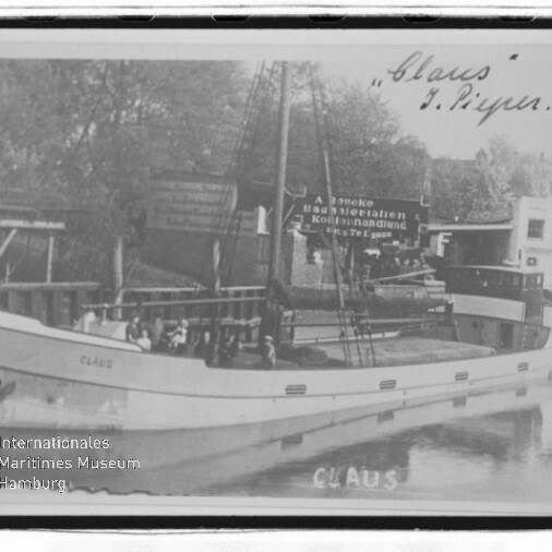 Schwarzweiß-Foto eines Frachtseglers Claus mit Mast und Namen am Bug, vertäut im Hafen; Museumswasserzeichen sichtbar.