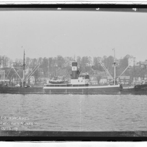 Frachtschiff vor Küste mit Häusern und Bäumen im Hintergrund, Schwarzweißaufnahme mit Museumslogo unten links.