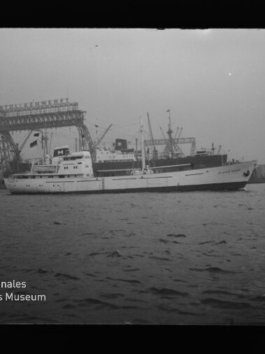 Weißes Schiff fährt im Hafen vor großen Werftkränen, ruhige See und Industriehintergrund mit Museumslogo unten links.