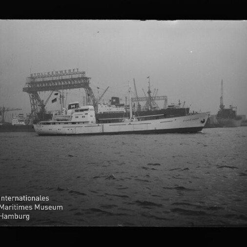 Weißes Schiff fährt im Hafen vor großen Werftkränen, ruhige See und Industriehintergrund mit Museumslogo unten links.
