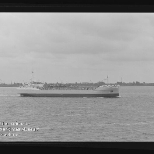 Frachtschiff in ruhiger See vor flachem Küstenhorizont, Schwarzweißaufnahme mit Museumswasserzeichen.