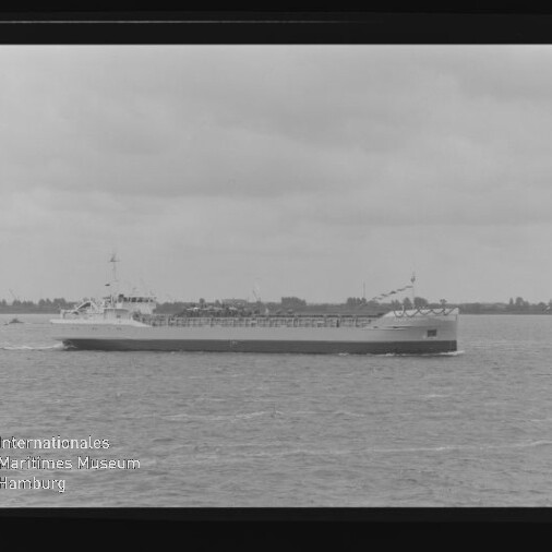 Langes Frachtschiff fährt auf ruhiger See vor flacher Küste unter bewölktem Himmel; Museumslogo unten links.