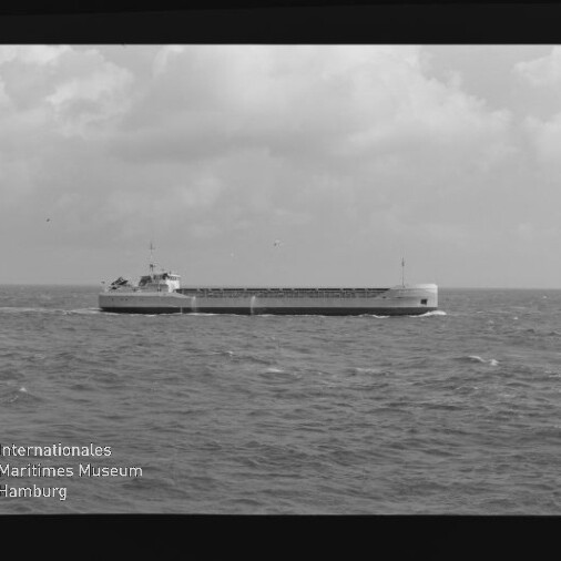 Schwarzweißfoto eines langen Frachtschiffs auf offener See unter Wolken; Wasserzeichen des Maritimen Museums.