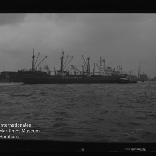 Großes Frachtschiff am Hafen mit Kränen im Hintergrund und Wasserfläche; links unten Logo des Internationalen Maritimen Museums Hamburg