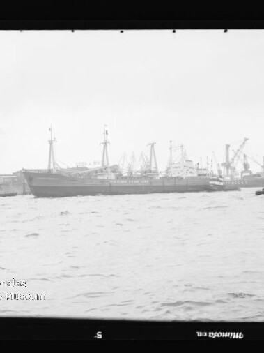Frachtschiff am Hafen mit mehreren Kranen und einem kleinen Boot im Vordergrund, Schwarzweißaufnahme