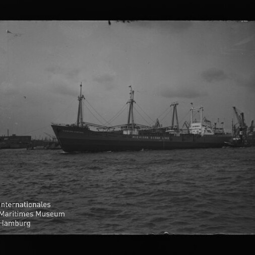 Großes Frachtschiff mit Ladebäumen und Masten im Hafen, Wasser im Vordergrund und Kräne im Hintergrund.