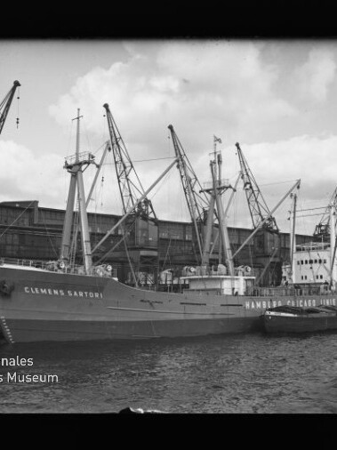 Schwarzweißfoto des Frachters Clemens Sartori am Kai, mehrere Hafenkräne und ein Schlepper neben dem Schiff.