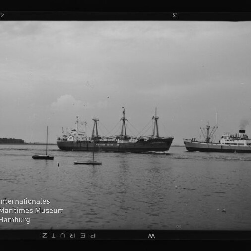 Zwei Frachtschiffe mit Ladekränen fahren nebeneinander auf ruhigem Wasser, kleine Segelboote im Vordergrund; schwarzweißfoto mit Museumsschrift.