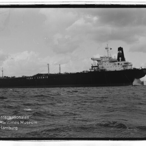 Schwarzweißaufnahme eines großen Tankers mit achterlichem Aufbauten, sichtbarer Bugwelle und bewölktem Himmel