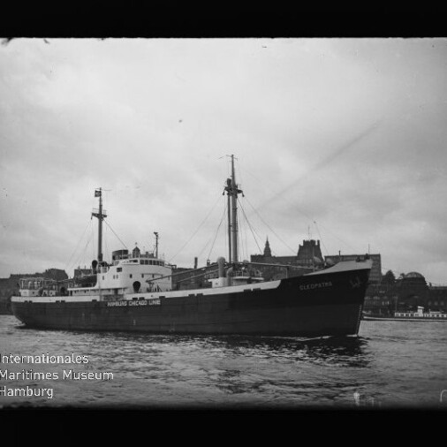 Frachtschiff mit dem Schriftzug Hamburg Chicago Line, Masten sichtbar, auf dem Wasser vor einer Stadtkulisse.