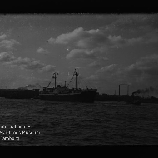 Großes Schiff mit geschmückten Masten vor Industriehafen und Wolkenhimmel; Logo Internationales Maritimes Museum Hamburg unten links