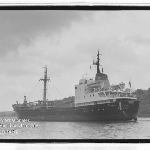 Großes Frachtschiff auf ruhigem Wasser vor bewaldeter Küste, Schwarzweiß-Foto mit Museums-Wasserzeichen.