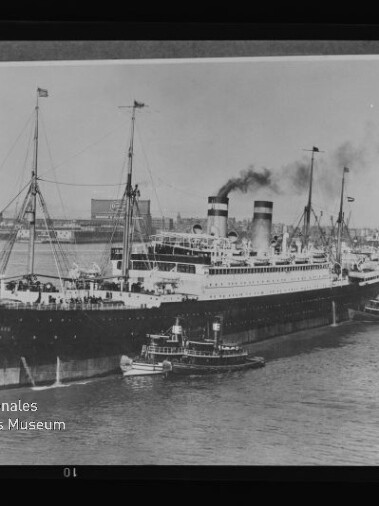 Großes historisches Passagierschiff mit zwei rauchenden Schornsteinen, begleitet von Schleppern im Hafen.