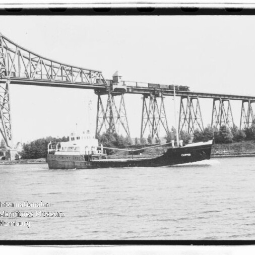 Frachtschiff 'Clipper' fährt auf dem Fluss vor einem hohen stählernen Eisenbahnviadukt mit Pylonen, Ufer mit Bäumen.