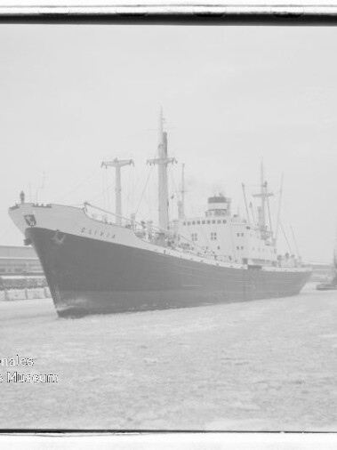 Großes Frachtschiff am Kai eines winterlichen Hafens mit Eis auf dem Wasser und Kränen im Hintergrund.