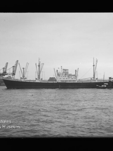 Frachtschiff mit Hafenkränen im Hintergrund und einem Schlepper an der Seite auf ruhigem Wasser.