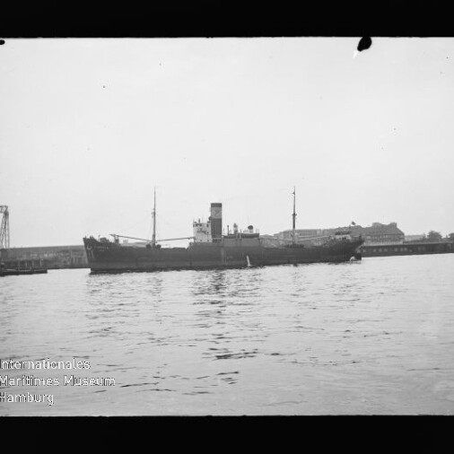 Frachtschiff mit mittigem Schornstein und zwei Masten vor Pier, spiegelnde ruhige Wasserfläche; Museumswasserzeichen links unten