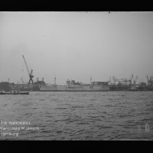 Großes Handelsschiff am Werftkai vor mehreren Hafenkränen, Wellengang im Vordergrund; kleines Boot links.