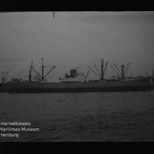 Seitenansicht eines Frachtschiffs mit Ladekränen am Hafen, ruhige See und Museumslogo links unten.