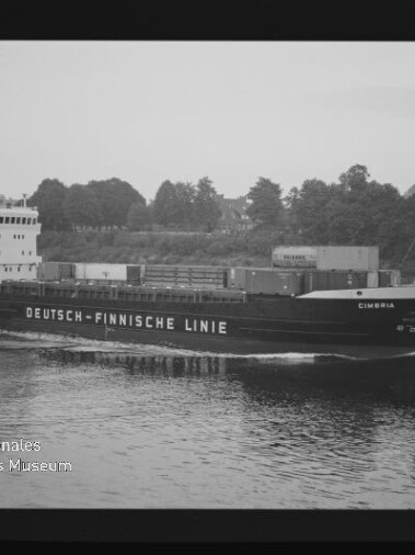 Frachtschiff Cimbria der Deutsch‑Finnischen Linie fährt auf ruhigem Wasser, Container an Deck, Baumufer im Hintergrund.