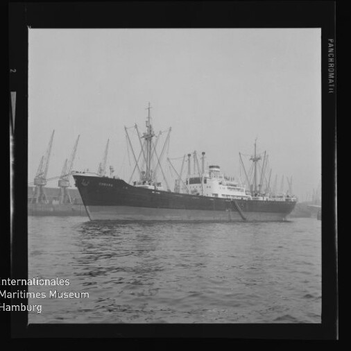 Schwarzweißfoto eines Frachtschiffs an einem Hafen mit Kränen im Hintergrund und Museumslogo unten links.