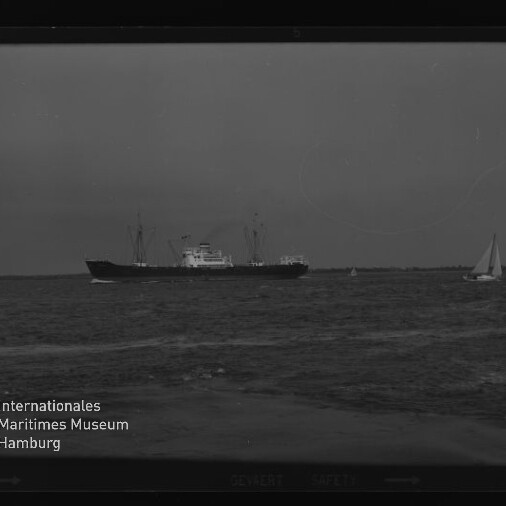 Frachtschiff mittig auf ruhiger See, kleines Segelboot rechts und Wasserzeichen Internationales Maritimes Museum links.