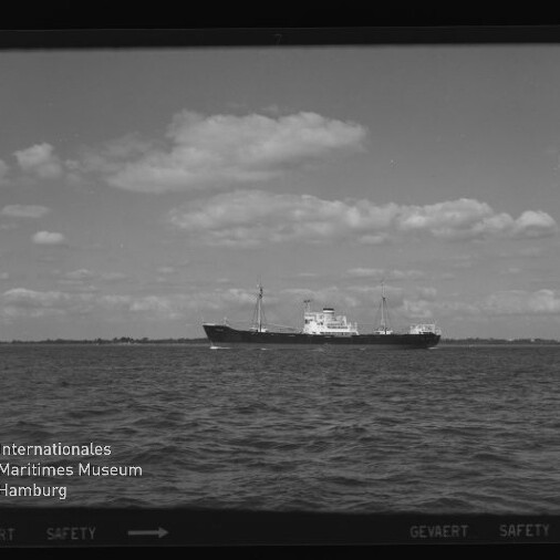 Frachtschiff auf ruhiger See vor flachem Küstenhorizont unter wolkigem Himmel; Logo des Maritimen Museums Hamburg sichtbar.