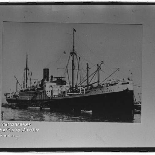 Schwarzweiß-Frachtdampfer mit Schornstein, Masten und Ladebäumen im Hafen; Museumswasserzeichen unten links.