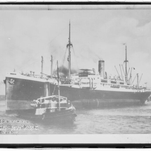 Großes Dampfschiff am Kai mit vorausfahrendem Schlepper, Rauch aus dem Schornstein und Hafenbauten im Hintergrund.