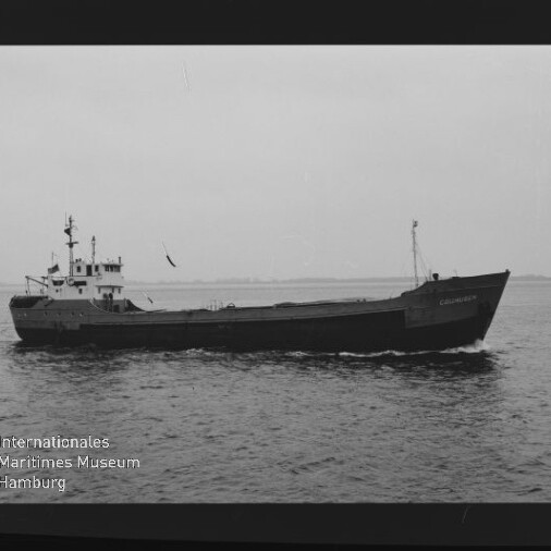 Schwarzweiß-Foto eines Frachtschiffs in Seitenansicht auf ruhiger See, mit Aufbauten achtern und niedrigem Ladedeck; deutlich sichtbarer Filmrand mit AGFAPAN 100-Aufdruck rechts und Museumswasserzeichen links unten.