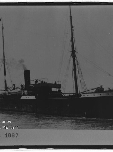 Schwarz-weiß-Foto des Dampfschiffs „Colonia“ (1887) in Seitenansicht im Hafen, mit einem Schornstein, zwei Masten, Flaggen und sichtbarem Wasserzeichen des Internationalen Maritimen Museums Hamburg.