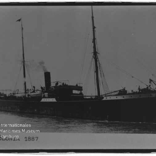 Schwarz-weiß-Foto des Dampfschiffs „Colonia“ (1887) in Seitenansicht im Hafen, mit einem Schornstein, zwei Masten, Flaggen und sichtbarem Wasserzeichen des Internationalen Maritimen Museums Hamburg.