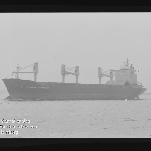 Graustufiges Foto eines Massengutfrachters mit drei großen Deckskränen und achterlicher Brücke, fahrend auf ruhiger See; unten links sichtbares Wasserzeichen des Internationalen Maritimen Museums Hamburg.