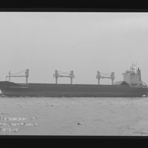 Schwarzweißfoto eines Frachtschiffs (Name sichtbar: Abraham Strength) mit drei Ladekränen und achterlichem Aufbauturm auf ruhiger See, umgeben vom Filmstreifenrand und mit Museumswasserzeichen unten links.
