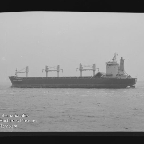 Schwarzweiß-Foto eines Massengutfrachters mit drei großen Ladekränen und achterlicher Aufbauten, fahrend auf ruhiger See; links Filmrand mit Schriftzug KODAK SAFETY FILM und unten links das Logo/ Wasserzeichen des Internationalen Maritimen Museums Hamburg.