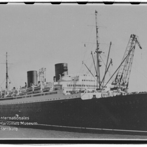 Schwarzweißaufnahme eines großen Passagierschiffs mit zwei hohen Schornsteinen am Kai, umgeben von Hafenkränen, im Vordergrund Wasser und links unten das Logo des Internationalen Maritimen Museums Hamburg